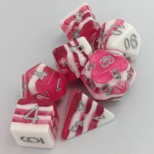 May include: A collection of seven polyhedral dice in pink and white with silver numerals. The dice have a marbled effect, with a combination of opaque and translucent areas. These dice are designed for tabletop role-playing games.