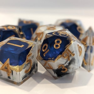 May include: A collection of polyhedral dice with a unique design. The dice feature a clear resin with a marbled white and gray effect, and deep blue accents. Gold-colored numbers and edges complete the look. Perfect for tabletop games.