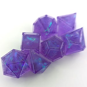Op de afbeelding: Een set van zeven paarse harsdobbelstenen met iriserende insluitsels. De dobbelstenen hebben verschillende polyhedrale vormen, waaronder een d20, d12, d10, d8, d6, d4 en een d10 percentieldobbelsteen. Elke dobbelsteen heeft witte nummering.