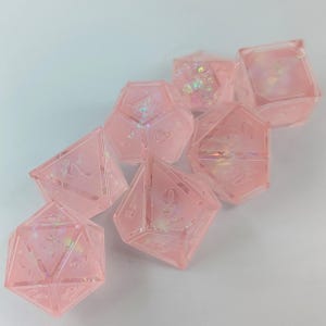 May include: A set of seven pink, translucent polyhedral dice. Each die has a different geometric shape and etched numbers. Some dice have iridescent glitter inside, creating a shimmering effect. The dice are designed for tabletop role-playing games.