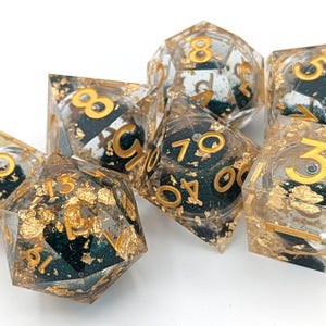 May include: Set of seven polyhedral dice. Each die is clear with gold numbers and gold leaf inclusions. The dice have a dark teal interior. The dice are for tabletop games.