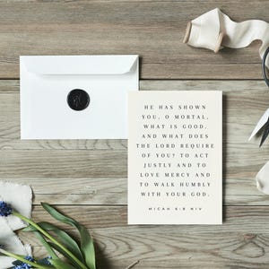 Puede incluir: Una tarjeta blanca con un sello de cera negra sobre una superficie de madera. La tarjeta tiene un fondo beige y un texto negro que dice "Él te ha mostrado, oh mortal, lo que es bueno. ¿Y qué requiere el Señor de ti? Actuar con justicia y amar la misericordia y caminar humildemente con tu Dios. Miqueas 6:8 NVI".