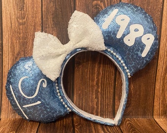 May include: Blue sequined mouse ear headband with a white bow. The ears have the text "1989" and "1st" in white. The headband is decorated with white pearls.