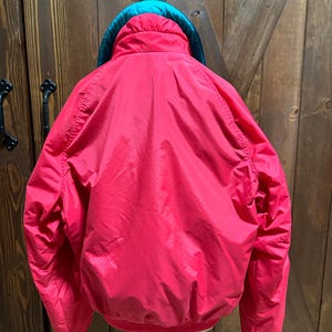 Puede incluir: Una chaqueta roja brillante con un cuello verde azulado y una parte superior negra. La chaqueta est&aacute; colgada en un soporte contra una puerta de madera. La chaqueta tiene una textura brillante y suave.