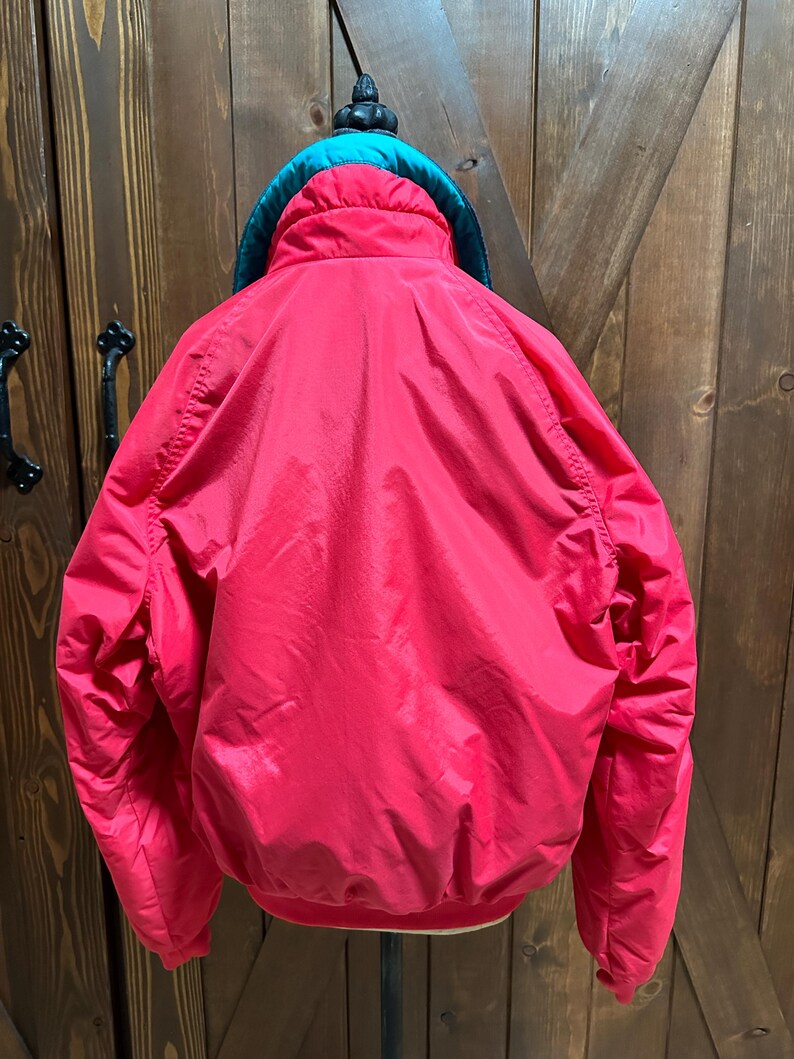 Puede incluir: Una chaqueta roja brillante con un cuello verde azulado y una parte superior negra. La chaqueta est&aacute; colgada en un soporte contra una puerta de madera. La chaqueta tiene una textura brillante y suave.