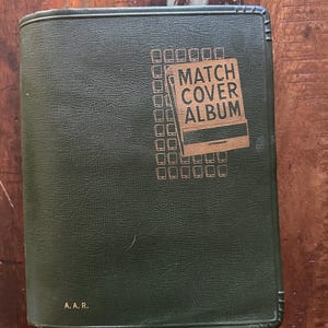 May include: A green match cover album with the words "MATCH COVER ALBUM" in gold lettering. The album has a textured surface and the initials "A. A. R." are printed at the bottom.