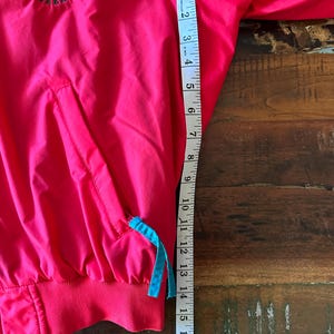 Puede incluir: Un cortavientos rosa brillante con el logotipo de Moosehead Canadian Lager en la parte superior izquierda. La chaqueta tiene un cord&oacute;n azul y un dobladillo acanalado rojo. La longitud de la chaqueta es de aproximadamente 45,7 cm.