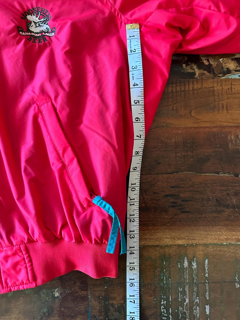 Puede incluir: Un cortavientos rosa brillante con el logotipo de Moosehead Canadian Lager en la parte superior izquierda. La chaqueta tiene un cord&oacute;n azul y un dobladillo acanalado rojo. La longitud de la chaqueta es de aproximadamente 45,7 cm.