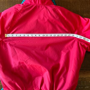 Puede incluir: Chaqueta rosa brillante con un acento turquesa y azul en el cuello. Una cinta m&eacute;trica muestra que el ancho de la chaqueta es de aproximadamente 68,5 cm. La chaqueta est&aacute; hecha de un material liso, posiblemente resistente al agua.