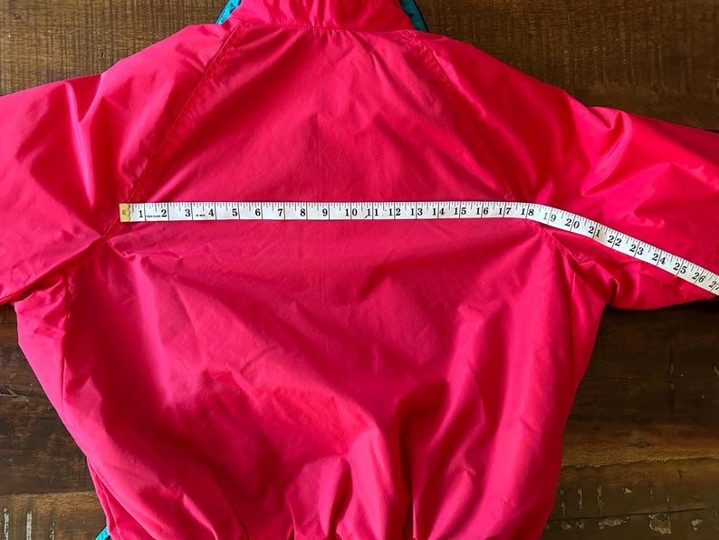 Puede incluir: Chaqueta rosa brillante con un acento turquesa y azul en el cuello. Una cinta m&eacute;trica muestra que el ancho de la chaqueta es de aproximadamente 68,5 cm. La chaqueta est&aacute; hecha de un material liso, posiblemente resistente al agua.