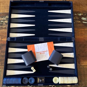 May include: Open backgammon set with a navy blue velvet interior, white and black playing spaces, and an orange booklet that reads "Backgammon Rules". Includes dice, doubling cube, and playing pieces.