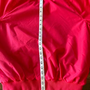 Puede incluir: Chaqueta rosa brillante con dobladillo y pu&ntilde;os de canal&eacute;. Una cinta m&eacute;trica muestra que la chaqueta mide aproximadamente 61 cm de largo. La chaqueta tiene cremallera y cuello alto. La chaqueta es de un material liso y ligero.