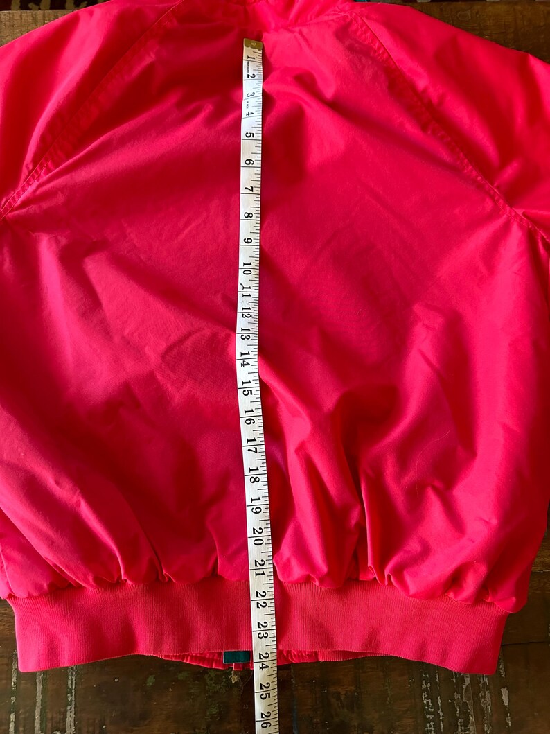 Puede incluir: Chaqueta rosa brillante con dobladillo y pu&ntilde;os de canal&eacute;. Una cinta m&eacute;trica muestra que la chaqueta mide aproximadamente 61 cm de largo. La chaqueta tiene cremallera y cuello alto. La chaqueta es de un material liso y ligero.