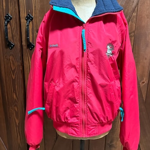 Puede incluir: Chaqueta roja Columbia con cuello forrado de forro polar azul y detalles turquesa. La chaqueta tiene cremallera completa, dos bolsillos y un peque&ntilde;o gr&aacute;fico bordado en la parte delantera. La chaqueta se muestra en un maniqu&iacute;.