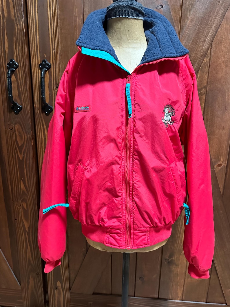 Puede incluir: Chaqueta roja Columbia con cuello forrado de forro polar azul y detalles turquesa. La chaqueta tiene cremallera completa, dos bolsillos y un peque&ntilde;o gr&aacute;fico bordado en la parte delantera. La chaqueta se muestra en un maniqu&iacute;.