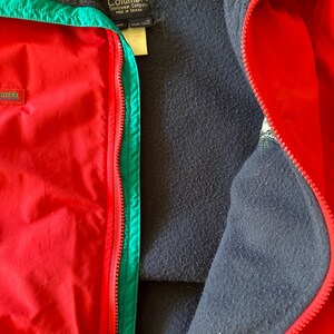 Puede incluir: Primer plano de una chaqueta Columbia roja con ribete verde e interior negro. La chaqueta tiene cierre de cremallera y un logotipo de Columbia en el pecho. La chaqueta est&aacute; hecha en Taiw&aacute;n.
