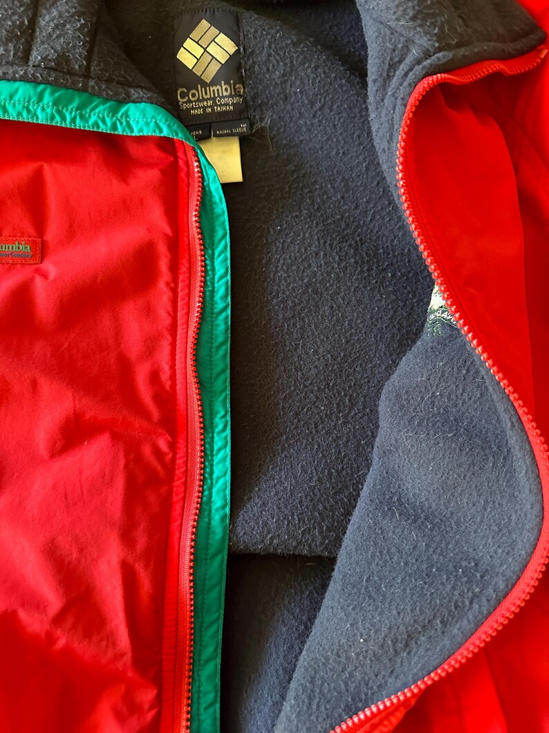 Puede incluir: Primer plano de una chaqueta Columbia roja con ribete verde e interior negro. La chaqueta tiene cierre de cremallera y un logotipo de Columbia en el pecho. La chaqueta est&aacute; hecha en Taiw&aacute;n.