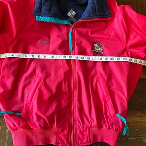 Puede incluir: Chaqueta Columbia rosa brillante con cuello de forro polar azul marino y detalles turquesa. La chaqueta tiene cierre de cremallera y un peque&ntilde;o logotipo bordado. La chaqueta mide aproximadamente 79 cm de ancho.