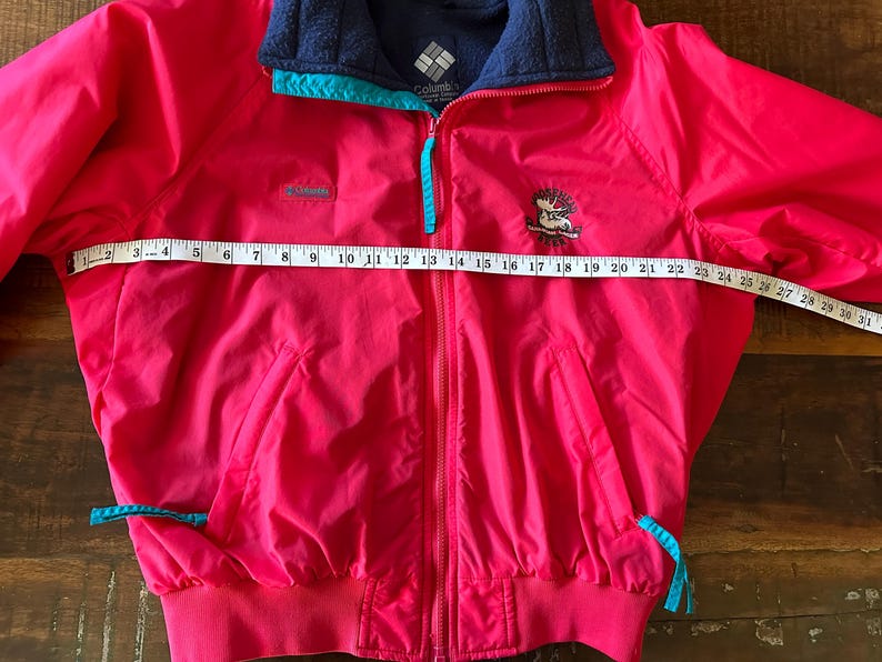 Puede incluir: Chaqueta Columbia rosa brillante con cuello de forro polar azul marino y detalles turquesa. La chaqueta tiene cierre de cremallera y un peque&ntilde;o logotipo bordado. La chaqueta mide aproximadamente 79 cm de ancho.