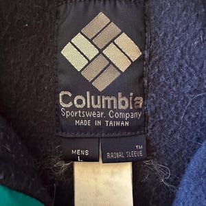 Puede incluir: Primer plano de una etiqueta azul marino de Columbia Sportswear Company. La etiqueta presenta el nombre y el logotipo de la marca, con texto que indica que est&aacute; fabricada en Taiw&aacute;n. Debajo hay etiquetas que dicen "MENS L" y "RADIAL SLEEVE".