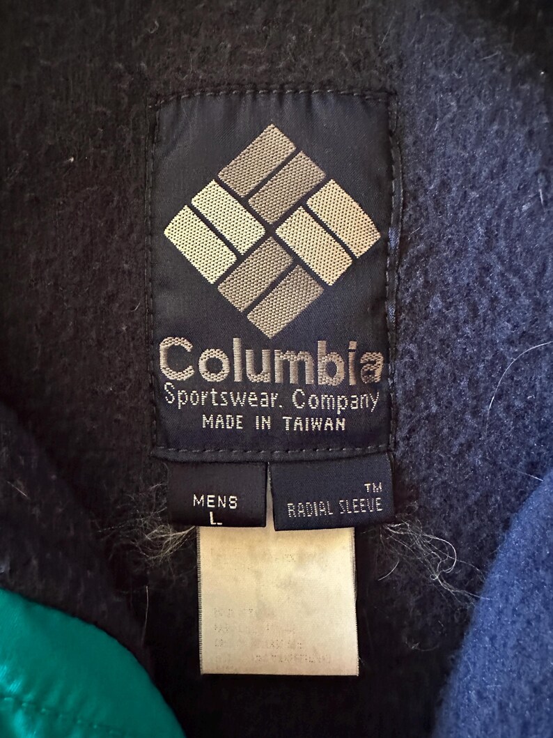 Puede incluir: Primer plano de una etiqueta azul marino de Columbia Sportswear Company. La etiqueta presenta el nombre y el logotipo de la marca, con texto que indica que est&aacute; fabricada en Taiw&aacute;n. Debajo hay etiquetas que dicen "MENS L" y "RADIAL SLEEVE".