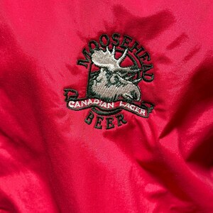Puede incluir: Primer plano de una chaqueta roja brillante con un logotipo bordado de Moosehead Canadian Lager Beer. El logotipo incluye una cabeza de alce, las palabras "Moosehead" y "Canadian Lager Beer" en una pancarta.