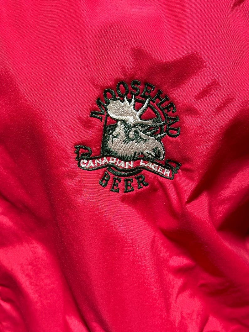 Puede incluir: Primer plano de una chaqueta roja brillante con un logotipo bordado de Moosehead Canadian Lager Beer. El logotipo incluye una cabeza de alce, las palabras "Moosehead" y "Canadian Lager Beer" en una pancarta.