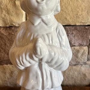 May include: A white ceramic figurine of a child with hands clasped in prayer. The figurine has a glossy finish and is dressed in a long-sleeved garment. The base of the figurine is chipped, revealing a terracotta color.