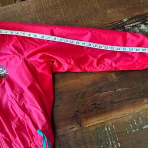 Puede incluir: Chaqueta rosa brillante con cuello azul y detalles turquesa. Una cinta m&eacute;trica muestra que la longitud de la manga es de aproximadamente 79 cm. La chaqueta tiene un peque&ntilde;o emblema bordado en el pecho. La chaqueta est&aacute; sobre una superficie de madera.