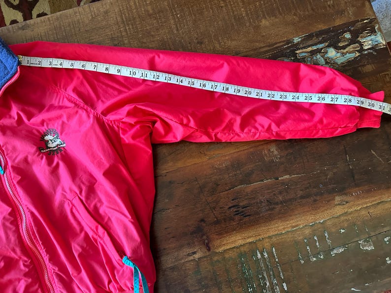Puede incluir: Chaqueta rosa brillante con cuello azul y detalles turquesa. Una cinta m&eacute;trica muestra que la longitud de la manga es de aproximadamente 79 cm. La chaqueta tiene un peque&ntilde;o emblema bordado en el pecho. La chaqueta est&aacute; sobre una superficie de madera.
