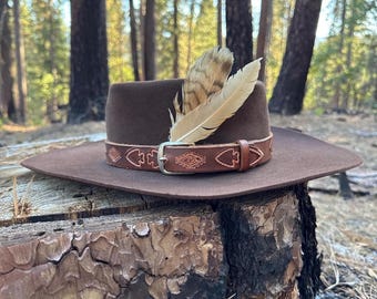 May include: A brown felt hat with a wide brim, adorned with a brown leather band featuring carved designs and a silver buckle. Two feathers are tucked into the hat's crown. The hat is set against a natural backdrop.