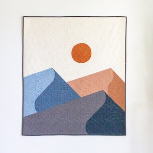 May include: A quilted wall hanging featuring a geometric design of mountains and a sun. The mountains are in shades of blue and grey, and the sun is orange. The background is white with a subtle pattern.