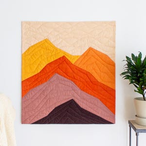Pode incluir: Uma tapeçaria de parede acolchoada com um design de cordilheira. As montanhas são representadas em tons de amarelo, laranja e roxo, com um céu pêssego claro. A peça é acolchoada à máquina com linhas diagonais.