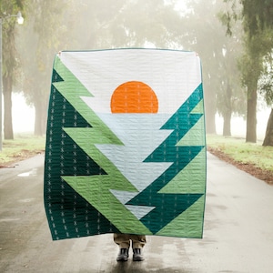May include: A green, white and orange geometric quilt with a design of a sunset behind a row of trees. The quilt is being held up in front of a person standing on a paved road.