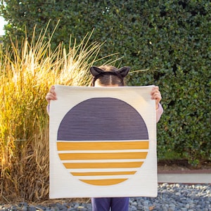 May include: A white quilted fabric square with a large circle in the center. The circle is divided in half, with the top half a dark purple and the bottom half a yellow with white stripes.