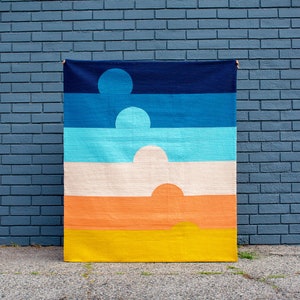 May include: A colourful woven tapestry with a geometric design of overlapping semi-circles in shades of blue, turquoise, cream, orange, and yellow. The tapestry is hanging against a blue brick wall.