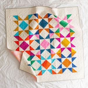 Puede incluir: Un edredón colorido con un patrón de estrellas geométricas. El edredón presenta un fondo color crema con estrellas vibrantes en tonos de rosa, naranja, azul, amarillo y verde. El edredón está hecho de tela y tiene una textura acolchada.