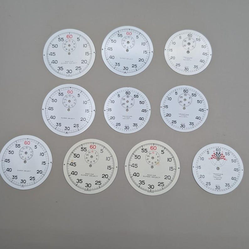May include: A collection of nine circular watch dials arranged in three rows. Each white dial features black numerical markings for seconds and minutes, with a smaller sub-dial at the top. Some dials display the text Racine Super Select or Yachting Timer.