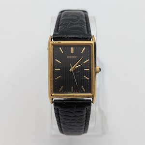 May include: A rectangular gold-toned Seiko watch with a black face and gold hands and markers. The watch has a black leather strap. The watch face has the word "SEIKO" printed on it.