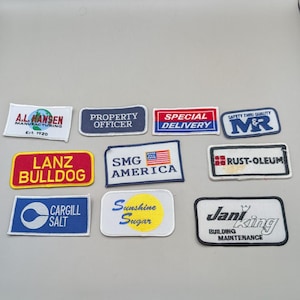 May include: A collection of eleven rectangular embroidered patches featuring various logos and text including A.L. Hansen, Property Officer, Special Delivery, M&R, Lanz Bulldog, SMG America with a flag, Rust-Oleum, Cargill Salt, Sunshine Sugar, and Jani-King.