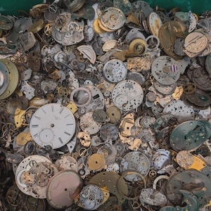May include: A collection of vintage watch parts, including dials, gears, and other internal components. The parts are in various shades of silver, gold, and bronze, with some showing signs of age and wear. The image showcases a variety of shapes and sizes, suggesting a wide range of watch models.