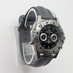 May include: A silver-toned Lucien Piccard watch with a black face and gray leather band. The watch features a moon phase indicator and day/date subdials. The watch has a round face with a rotating bezel and is displayed on a clear stand.