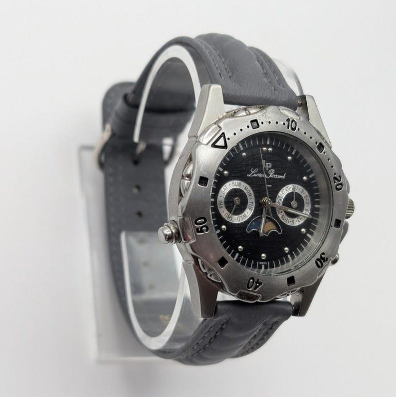 May include: A silver-toned Lucien Piccard watch with a black face and gray leather band. The watch features a moon phase indicator and day/date subdials. The watch has a round face with a rotating bezel and is displayed on a clear stand.