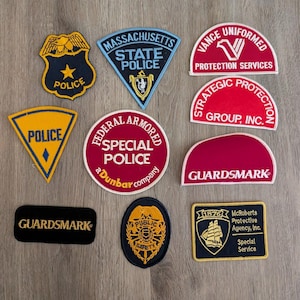 May include: A collection of ten embroidered patches featuring various security and police emblems. The patches include text such as Massachusetts State Police, Vance Uniformed Protection Services, Strategic Protection Group, Inc., Federal Armored Special Police, Guardsmark, Public Safety, and McRoberts Protective Agency, Inc. in various shapes and colours.
