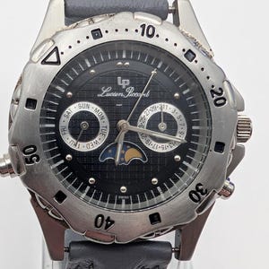 May include: A Lucien Piccard men's watch with a black dial, silver-tone bezel, and gray leather strap. The watch features day, date, and moon phase complications. The watch case is approximately 40mm in diameter.