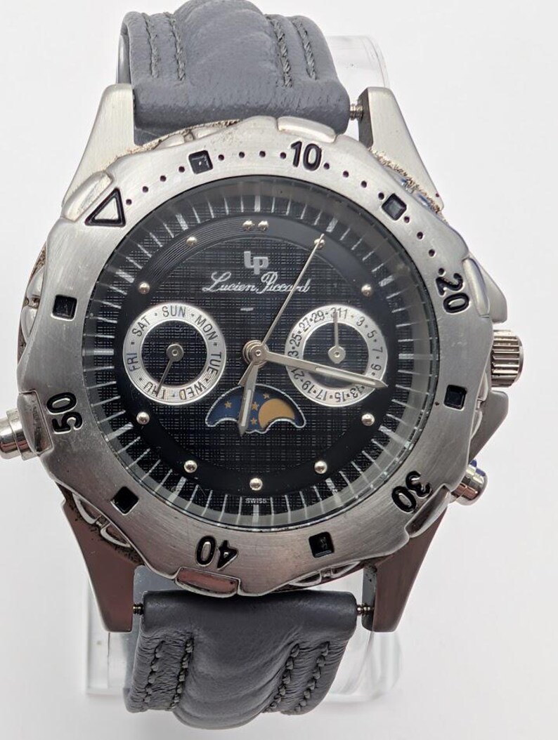 May include: A Lucien Piccard men's watch with a black dial, silver-tone bezel, and gray leather strap. The watch features day, date, and moon phase complications. The watch case is approximately 40mm in diameter.