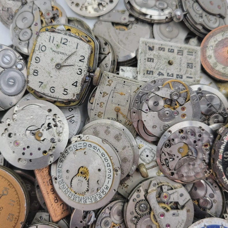 May include: A collection of vintage watch parts, including gears, dials, and movements. The image features a variety of shapes and sizes, with visible numbers and hands on some dials. An Ingersoll watch face is prominent, showing the time.
