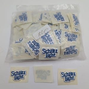 May include: A clear plastic bag containing 100 pieces of small, rectangular white patches with the blue embroidered text "Schlitz Light" in a stylized font. Three individual patches are placed in front of the bag on a white surface.