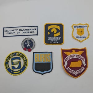 May include: A collection of six embroidered security patches featuring various shapes and designs, including rectangular, circular, and shield-like forms with text such as Security Management Group of America, Pinkerton, Burns, and Pasco County Security Patrol.