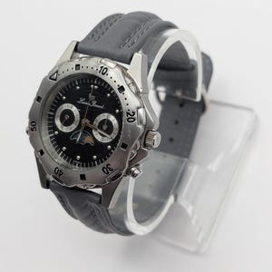 May include: A silver-toned wristwatch with a black face and gray leather band. The watch features multiple dials and a moon phase indicator. The bezel has a textured design, and the watch is displayed on a clear stand.
